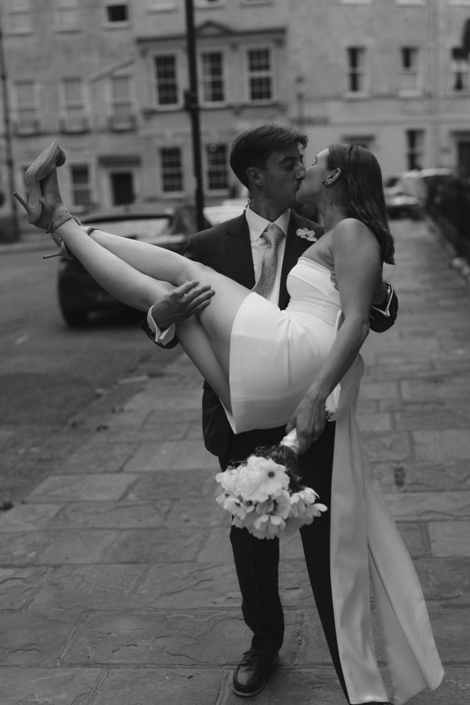 Groom lifts bride into his arms for a romantic kiss in Bath city centre after their wedding